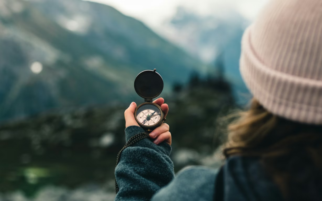 A woman stands back on in a mountain range (out of focus in the far distance) holding a compass. She's wearing a pink toque over her brown hair and a dark blue sweatshirt.